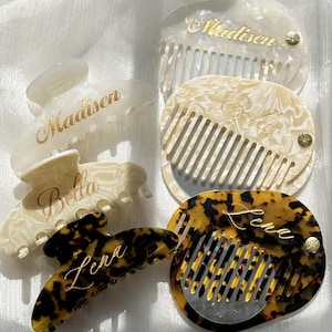 May include: A selection of personalised hair accessories, including hair claws and combs. The hair claws are in cream and tortoiseshell patterns, with the names "Madisen", "Bella", and "Lena" in gold script. The combs are cream and tortoiseshell, also with gold lettering.