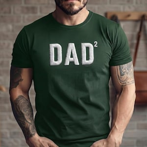 May include: Dark green t-shirt with the word "DAD²" embroidered in white. The shirt has a crew neck and short sleeves. The image shows a person wearing the shirt.