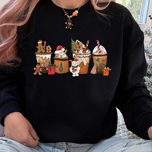 May include: Black sweatshirt featuring a festive holiday design. The graphic depicts several mugs of hot chocolate and gingerbread characters. The mugs are decorated with whipped cream, candy canes, and Christmas trees. A perfect gift for the holiday season.