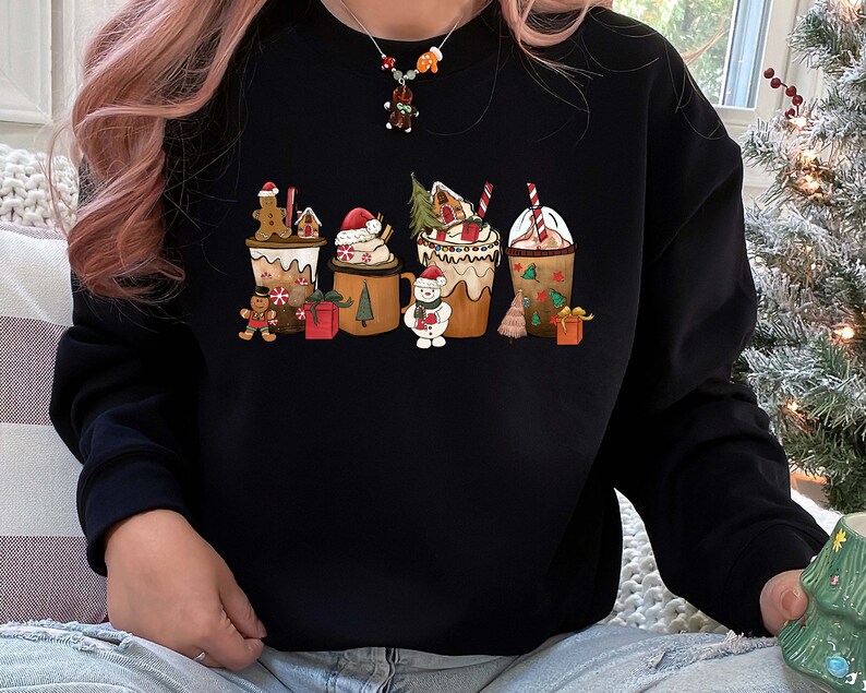 May include: Black sweatshirt featuring a festive holiday design. The graphic depicts several mugs of hot chocolate and gingerbread characters. The mugs are decorated with whipped cream, candy canes, and Christmas trees. A perfect gift for the holiday season.