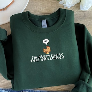 Pu&ograve; includere: Una felpa verde scuro con un'immagine di pollo pixelata e il testo "Sto iniziando a sentirmi esausto."