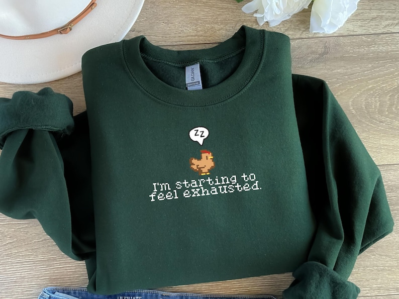 Pu&ograve; includere: Una felpa verde scuro con un'immagine di pollo pixelata e il testo "Sto iniziando a sentirmi esausto."