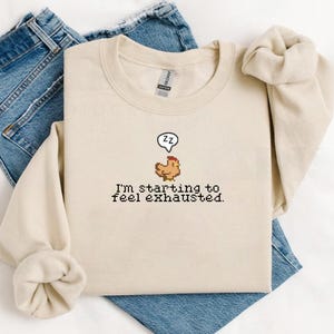 Pu&ograve; includere: Una felpa beige con un'immagine pixelata di un pollo con un fumetto che dice "zz" e il testo "Sto iniziando a sentirmi esausto."