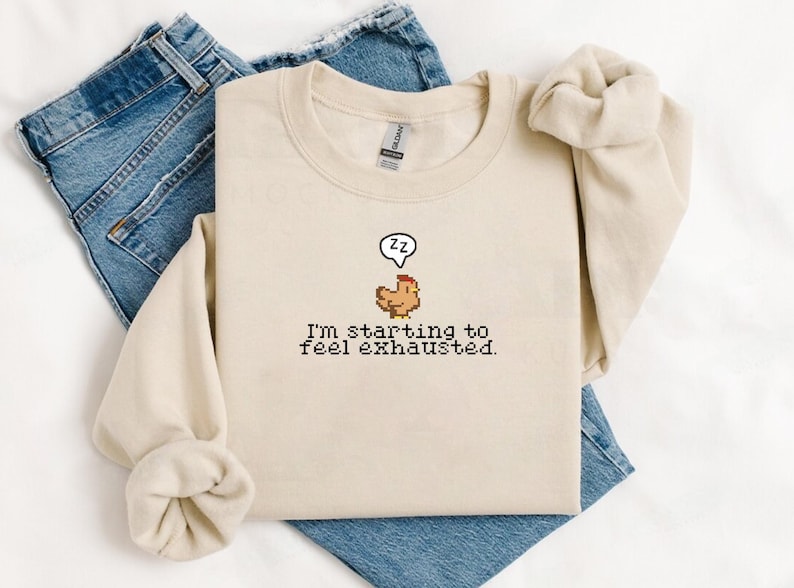 Pu&ograve; includere: Una felpa beige con un'immagine pixelata di un pollo con un fumetto che dice "zz" e il testo "Sto iniziando a sentirmi esausto."