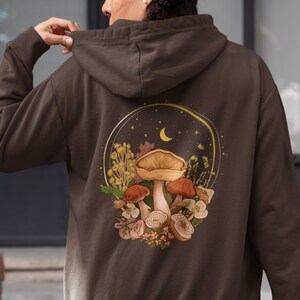 Peut inclure: Un sweat &agrave; capuche marron avec un motif de champignons, de fleurs et d'un croissant de lune dans un cercle au dos.