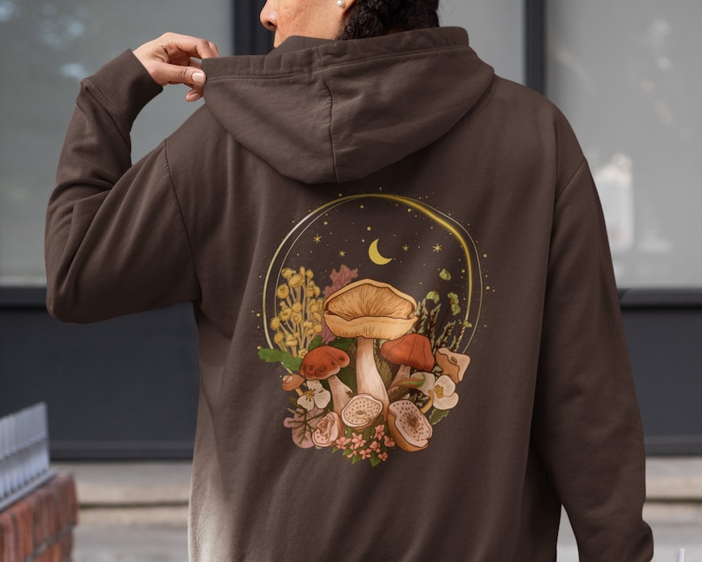Peut inclure: Un sweat &agrave; capuche marron avec un motif de champignons, de fleurs et d'un croissant de lune dans un cercle au dos.