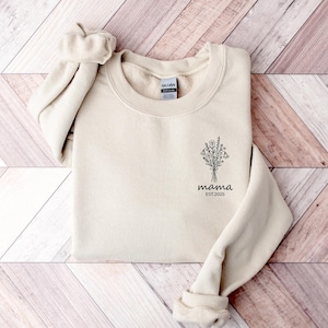 Könnte beinhalten: Ein cremefarbenes Sweatshirt mit Rundhalsausschnitt. Auf der Vorderseite befindet sich ein schwarzes Blumenstrauß-Design mit dem Wort "mama" und dem Jahr "EST.2025" darunter. Das Sweatshirt liegt auf einer Holzoberfläche.