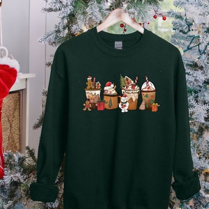 May include: Dark green sweatshirt featuring a festive Christmas-themed design. The graphic depicts five coffee cup-shaped designs, each adorned with gingerbread men, Santa hats, and other holiday elements. The sweatshirt is hanging on a wooden hanger.