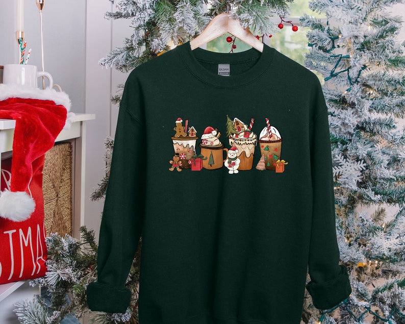 May include: Dark green sweatshirt featuring a festive Christmas-themed design. The graphic depicts five coffee cup-shaped designs, each adorned with gingerbread men, Santa hats, and other holiday elements. The sweatshirt is hanging on a wooden hanger.