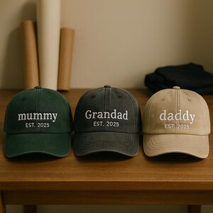 May include: Three baseball caps in green, gray, and tan. Each cap has white embroidered text: "mummy", "Grandad", and "daddy", with "EST. 2025" below. The caps are on a wooden surface.