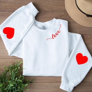 Può includere: Felpa bianca con un cuore rosso su ogni manica e la parola "love" in corsivo rosso sul petto. La felpa è a girocollo. Le maniche hanno un cuore rosso su ogni manica.