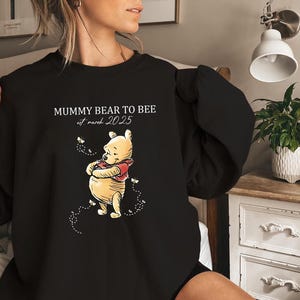 May include: A black crewneck sweatshirt featuring a graphic of Winnie the Pooh surrounded by small bees. The text reads MUMMY BEAR TO BEE and est. march 2025 in white lettering. The design is centred on the chest of the long-sleeved garment.