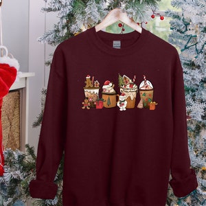 May include: Burgundy sweatshirt featuring a festive Christmas-themed design. The graphic includes several decorated coffee cups with gingerbread men, Santa hats, and other holiday elements. The sweatshirt is hanging on a wooden hanger.