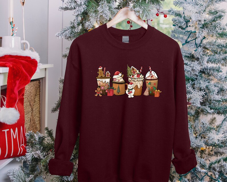 May include: Burgundy sweatshirt featuring a festive Christmas-themed design. The graphic includes several decorated coffee cups with gingerbread men, Santa hats, and other holiday elements. The sweatshirt is hanging on a wooden hanger.