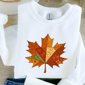 Op de afbeelding: Witte crewneck sweatshirt met een groot, kleurrijk esdoornbladontwerp. Het blad is samengesteld uit verschillende stofpatronen in tinten oranje, bruin en groen, wat een herfstthema oproept. De sweatshirt is gemaakt van een zacht materiaal.