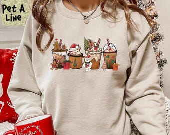 May include: A beige sweatshirt featuring a festive graphic of five holiday-themed coffee cups. Each cup is decorated with gingerbread men, Santa hats, and other Christmas elements. The text "Pet A Line" is visible in the upper left corner.