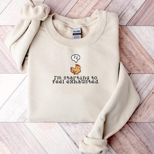 Pu&ograve; includere: Una felpa beige con una grafica di pollo pixelata e il testo "Sto iniziando a sentirmi esausto."