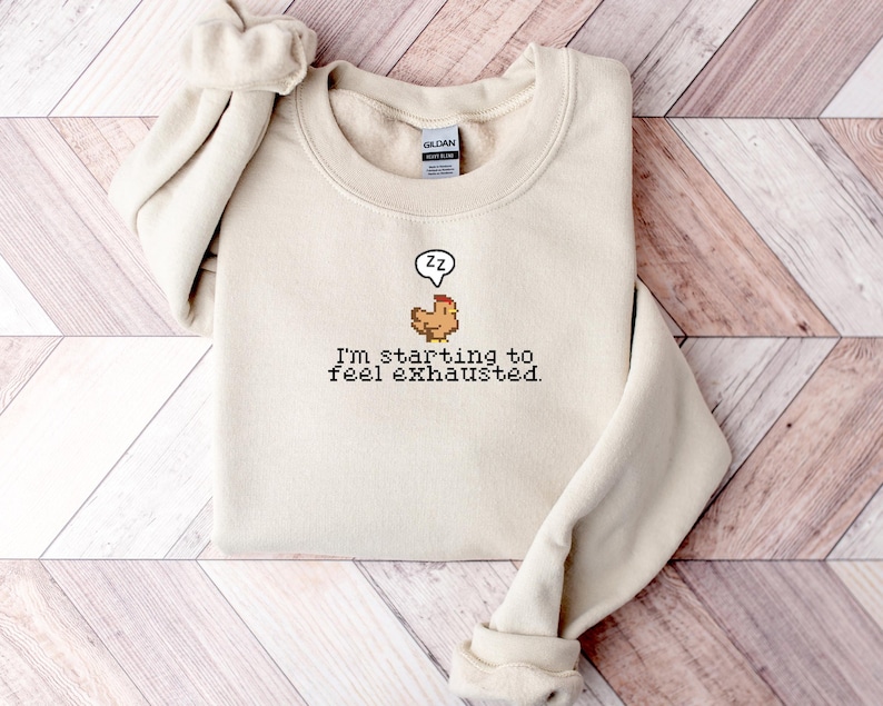 Pu&ograve; includere: Una felpa beige con una grafica di pollo pixelata e il testo "Sto iniziando a sentirmi esausto."