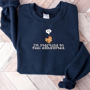 Pu&ograve; includere: Una felpa blu navy con un designo ricamato bianco di un pollo con un fumetto che dice "zz" e il testo "Sto iniziando a sentirmi esausto."