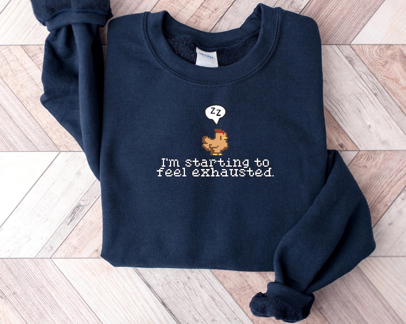 Pu&ograve; includere: Una felpa blu navy con un designo ricamato bianco di un pollo con un fumetto che dice "zz" e il testo "Sto iniziando a sentirmi esausto."