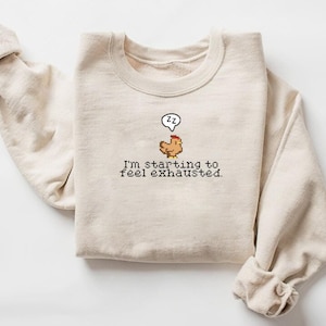 Pu&ograve; includere: Una felpa beige con un'immagine di pollo pixelata e il testo "Sto iniziando a sentirmi esausto."