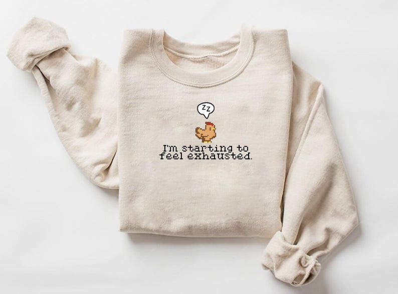 Pu&ograve; includere: Una felpa beige con un'immagine di pollo pixelata e il testo "Sto iniziando a sentirmi esausto."