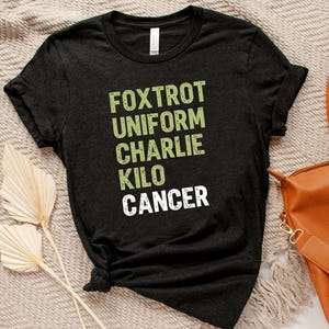 May include: A black t-shirt with the words "Foxtrot Uniform Charlie Kilo Cancer" printed in green and white. The shirt is folded and lies on a textured surface, with a brown bag and dried plants in the background.