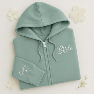May include: A sage green zip-up hoodie with the word "Bride" and the date "28.6.24" printed in white script. The left sleeve cuff has the name "Sophia" and a heart printed in white. The hoodie has a hood and a front zipper.