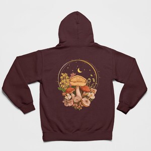 Peut inclure: Un sweat &agrave; capuche marron avec un motif de champignons et de fleurs dans un cercle dor&eacute; avec une lune croissante et des &eacute;toiles.