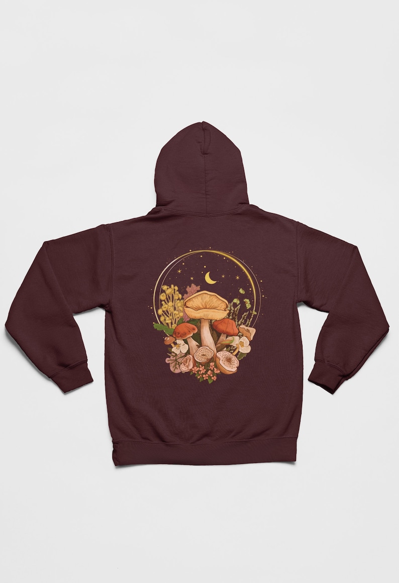 Peut inclure: Un sweat &agrave; capuche marron avec un motif de champignons et de fleurs dans un cercle dor&eacute; avec une lune croissante et des &eacute;toiles.