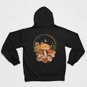Peut inclure: Sweat-shirt &agrave; capuche noir avec un motif de champignons et de fleurs dans un cercle dor&eacute; avec une lune croissante et des &eacute;toiles.