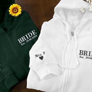 Può includere: Due felpe con zip, una verde e una bianca, con la parola "BRIDE" ricamata sul davanti. La felpa verde ha "Mrs. B" ricamata sulla manica, e la felpa bianca ha "Mrs. F" ricamata sulla manica. Entrambe le felpe hanno la data "Est. 29.06.2024" ricamata sotto la parola "BRIDE".