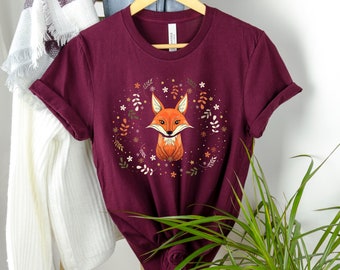 Puede incluir: Camiseta color burdeos con una ilustración de un zorro adorable rodeado de detalles florales. El zorro es naranja y blanco con la nariz y los ojos negros.