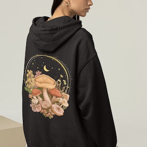 Peut inclure: Sweat-shirt &agrave; capuche noir avec un imprim&eacute; graphique de champignons, de fleurs et d'un croissant de lune au dos. Le graphique est entour&eacute; d'un cercle dor&eacute; et d'&eacute;toiles.