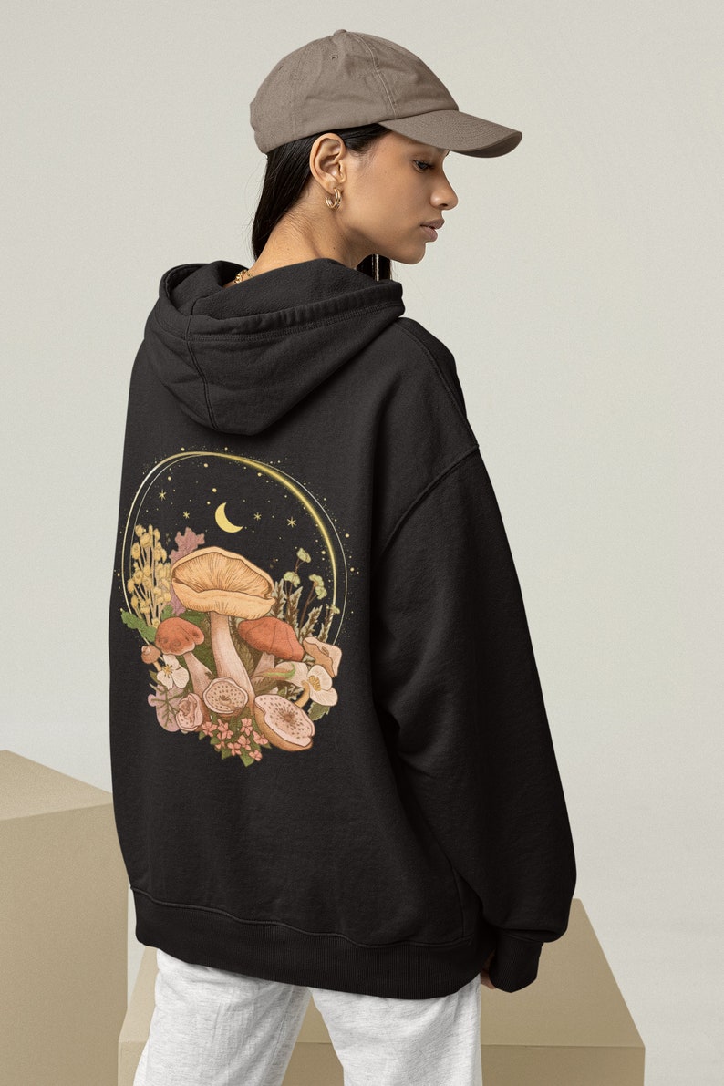 Peut inclure: Sweat-shirt &agrave; capuche noir avec un imprim&eacute; graphique de champignons, de fleurs et d'un croissant de lune au dos. Le graphique est entour&eacute; d'un cercle dor&eacute; et d'&eacute;toiles.