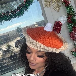 May include: A crocheted pie-shaped hat with an orange top and a tan crust. The hat has a white dollop of whipped cream on top. The image includes the text "@mariemadeit__".