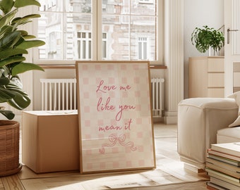 May include: A framed print with a pink and white checkered background. The text "Love me like you mean it" is written in pink cursive font. A pink ribbon with a bow is at the bottom of the print.