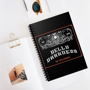 May include: Black spiral-bound notebook with the text "HELLO DARKNESS MY OLD FRIEND" in white gothic lettering. The cover features a design with two skulls, a sun, and bats, framed in orange.