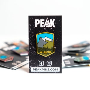 May include: A black and white enamel pin with a yellow border featuring a mountain range with a blue sky and white snow. The pin says "Ha Ling Peak" and "PEAK" in white text on a black background. The pin is on a black card with social media icons and the website "PEAKPINS.COM".