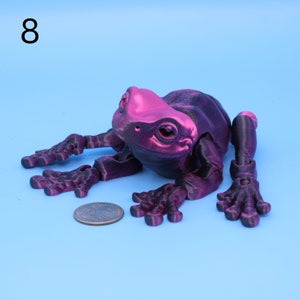 May include: A 3D printed articulated frog figurine with a black and pink metallic finish. The frog is posed sitting with its legs outstretched.