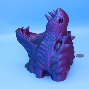 May include: A 3D-printed, purple and blue dragon sculpture with an open mouth and sharp teeth. The dragon has textured scales and a detailed design. The sculpture is on a light blue background.