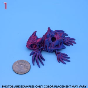 May include: A 3D printed, purple and red, skeletal dragon figurine. The dragon is lying on its side with its wings spread out. The figurine is about the size of a US quarter.