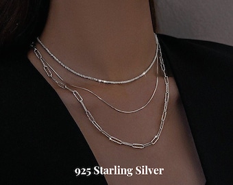 Op de afbeelding: Een set van drie zilveren kettingen, waaronder een delicate ketting met kleine steentjes, een dunne ketting en een dikkere ketting met grotere schakels. De kettingen zijn gelaagd op de nek van een model. De tekst "925 Starling Silver A+B+C Chain Set" wordt weergegeven onder de kettingen.