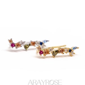 May include: A pair of gold ear cuffs with a row of colorful gemstones. The gemstones are in various shapes and sizes, including round, oval, and triangular. The ear cuffs are a delicate and stylish accessory.