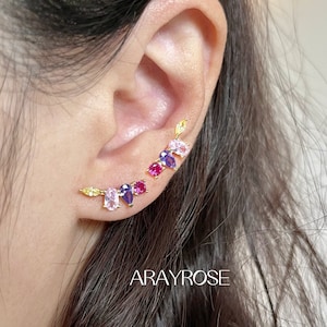 May include: A pair of gold ear cuffs with colorful gemstones. The ear cuffs feature a row of pink, yellow, blue, and red gemstones.