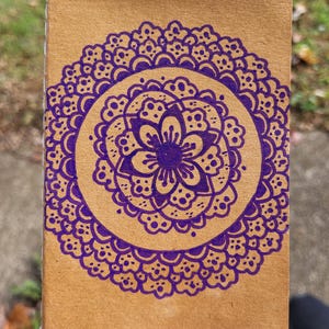 May include: A brown paper notebook with a purple mandala design. The mandala features a flower in the centre surrounded by intricate patterns.