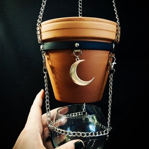 May include: A terracotta hanging planter with a black leather strap and a crescent moon charm. The planter is suspended from a silver chain and is hanging over a clear glass bowl.
