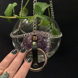 Puede incluir: Un macetero colgante de vidrio en forma de esfera con una cadena plateada y una correa de cuero negro con un anillo plateado. El macetero está lleno de cristales de amatista y una planta verde está creciendo dentro.