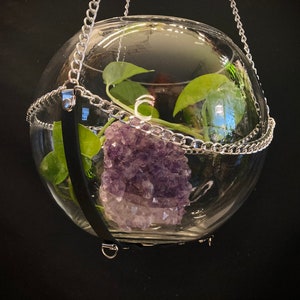May include: A glass orb hanging from a silver chain with a large amethyst crystal and a green plant inside.