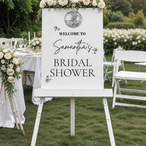 Könnte beinhalten: Ein weißes Staffeleischild mit den Worten "Welcome to Samantha's Bridal Shower" in schwarzer Schrift. Das Schild ist mit einer Discokugel und Blumengestecken verziert. Das Schild steht im Freien auf einer Rasenfläche.